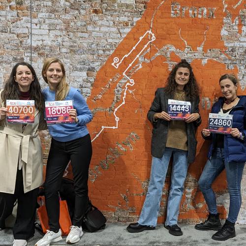 Four club members holding their race bibs in front of the course map mural at the TCS New York City Marathon expo
