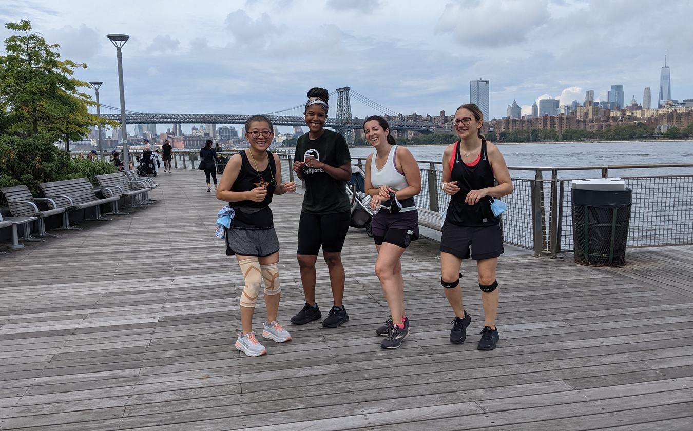 Four club members on a waterfront boardwalk during a group run, with the Williamsburg Bridge and Manhattan skyline behind them